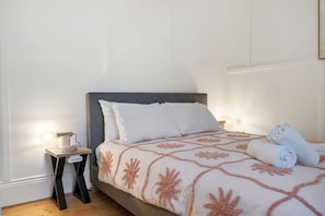 Ein ordentlich bezogenes Bett mit einem gemusterten Überwurf, ein Nachttisch mit Lampe und gerollte Handtücher auf dem Boden.