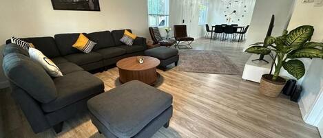 A modern living room with a sectional sofa, ottoman, and coffee table. There is a dining area with a table and chairs in the background.