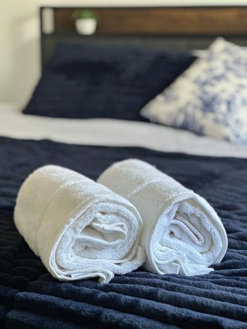 A neatly made bed with a dark blue quilt, white pillows, and a rolled white towel on the bed.