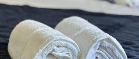 A neatly made bed with a dark blue quilt, white pillows, and a rolled white towel on the bed.