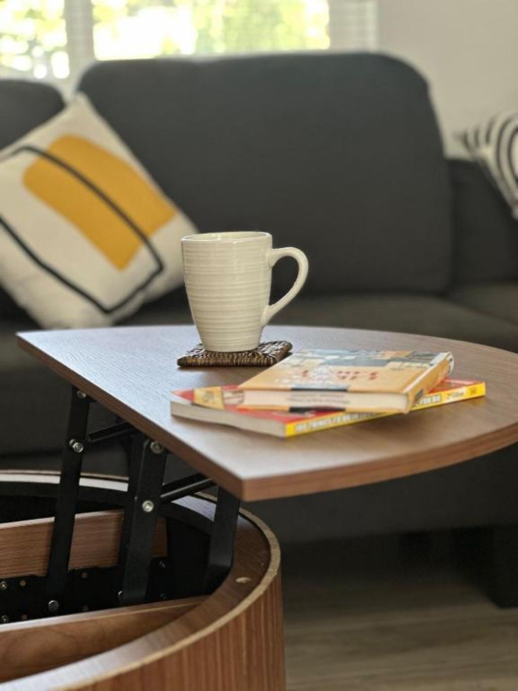 A small wooden table with a white mug and a stack of books on it, placed in front of a grey sofa with a yellow and black pillow.