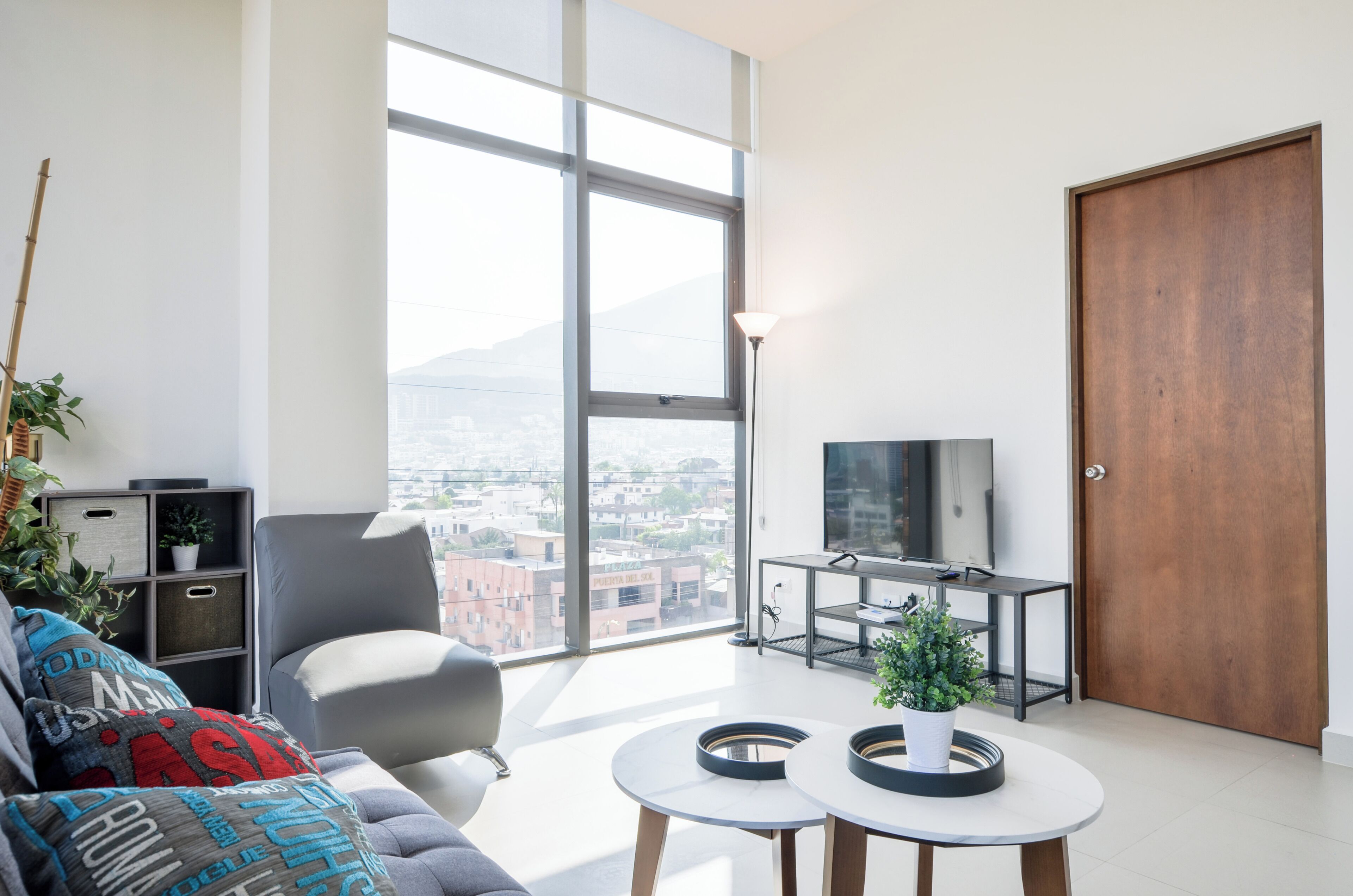 A modern living room with a large window, a sofa, a TV on a stand, and a door.