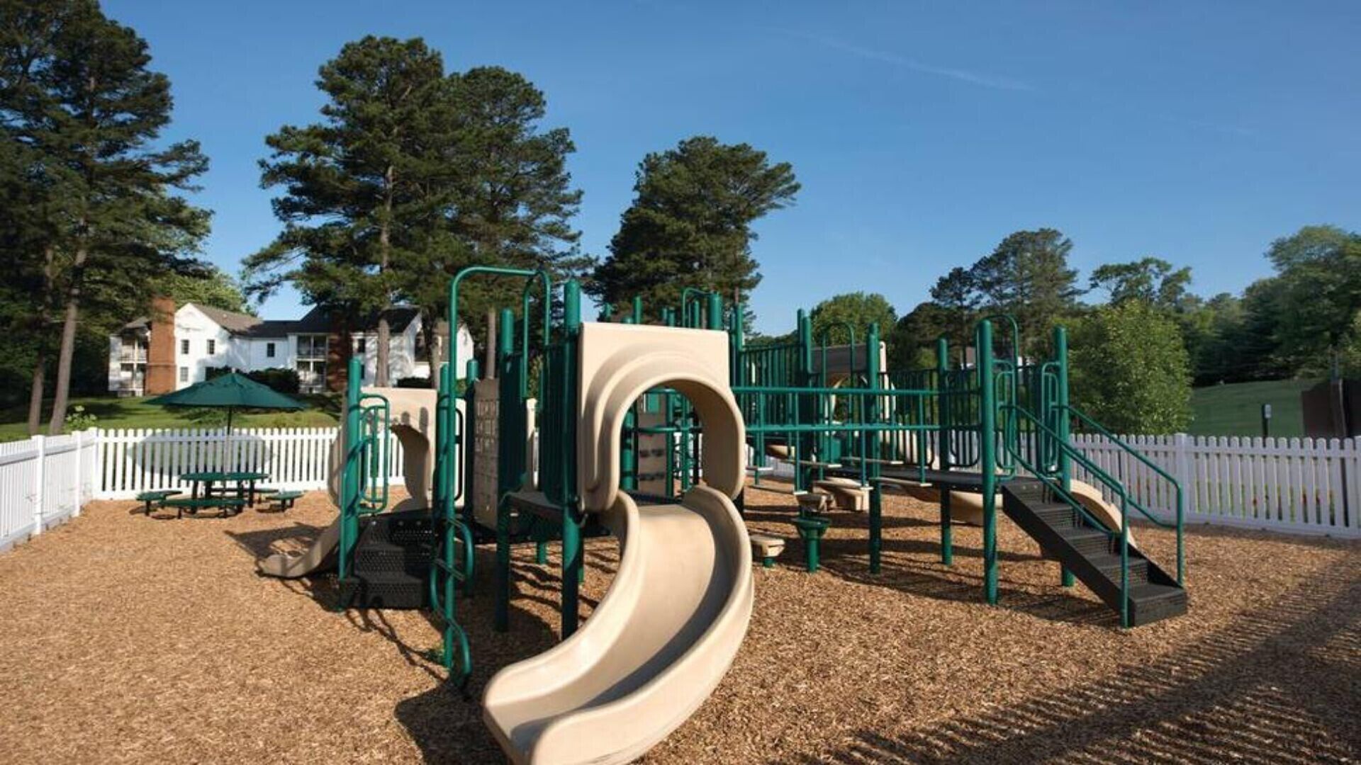 A playground with a slide and climbing structures, surrounded by a white picket fence and trees.