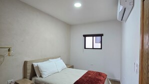 A bedroom with a bed, bedside table, nightstand, wall-mounted air conditioning unit, window, and a door. - New centrally located apartment with shared balcony. (Chihuahua)