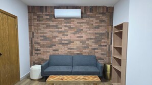 A living room with a blue sofa, a wooden coffee table, a brick-patterned wall, a white air conditioner, and a wooden bookshelf. - New centrally located apartment with shared balcony. (Chihuahua)