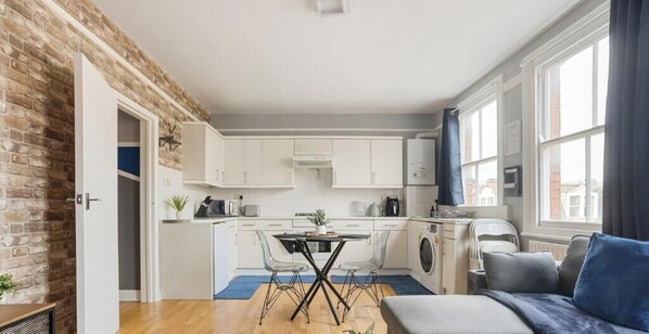 A modern kitchen with white cabinets, a dining table with chairs, a washing machine, and a large window. - Relaxing 2BR Flat|Near Stores|Near Station|ST (Greater London)