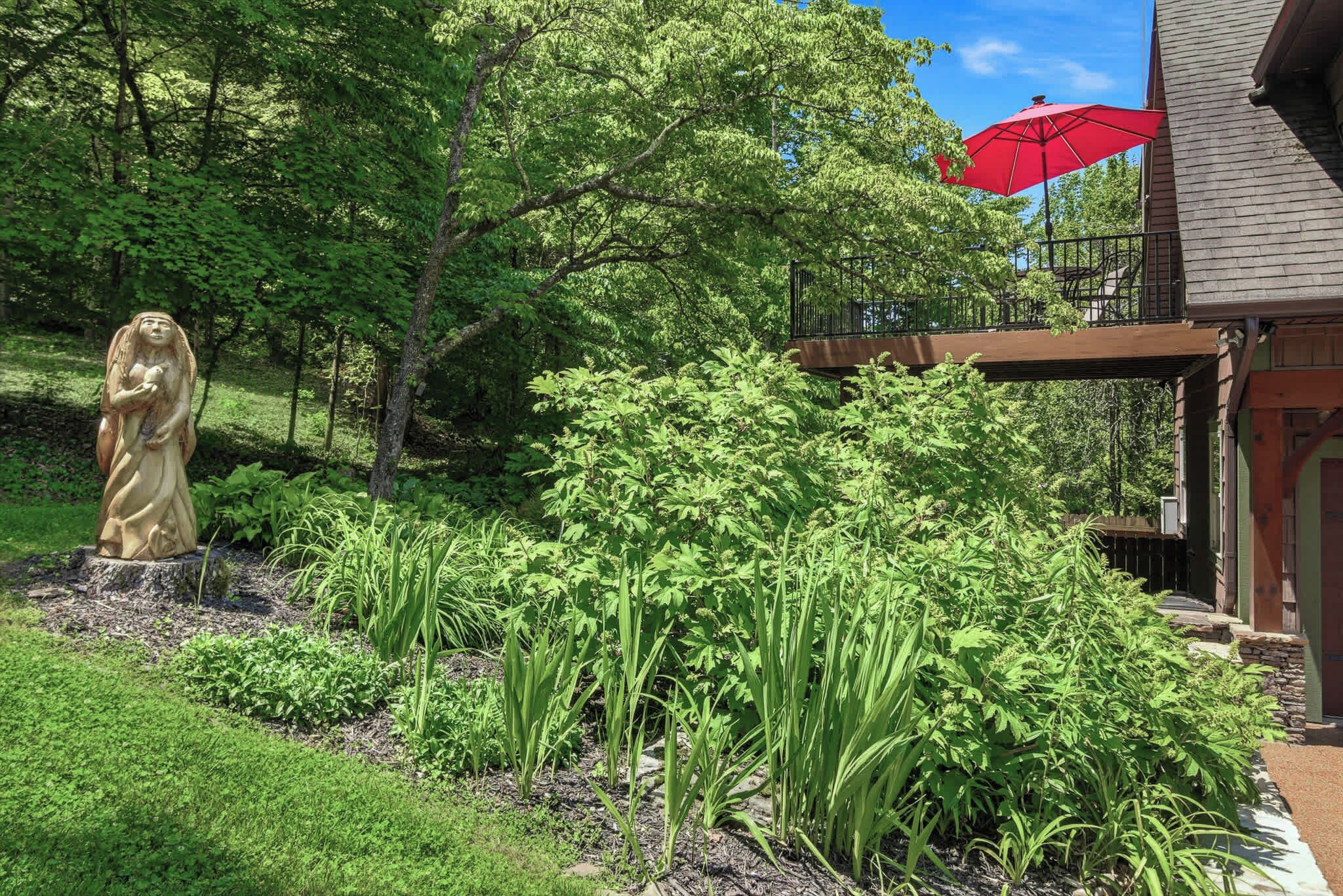 A garden with a statue, lush greenery, and a building with a balcony and red umbrella.
