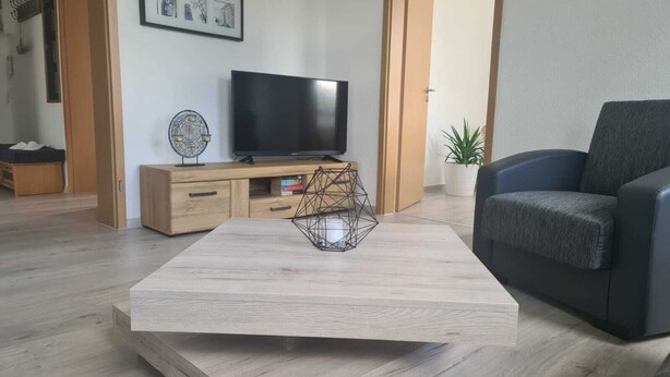 Ett modernt vardagsrum med ett träbord, en svart TV placerad på ett träställ, en grå soffa och en dekorativ skulptur gjord av metalltråd.