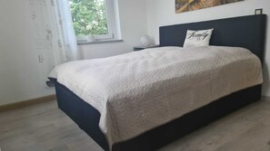 Une chambre à coucher avec un lit, une table de chevet, une fenêtre donnant sur des arbres et un mur orné d’un tableau.
