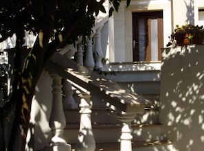 Una escalera con barandilla blanca que lleva a una puerta con ventana, rodeada de follaje.