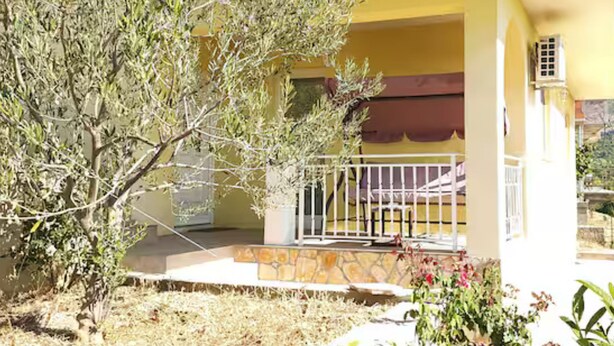Un edificio amarillo con cimientos de piedra, barandal blanco y un pequeño jardín con plantas y flores.