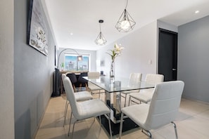 A modern dining area with a glass table surrounded by white chairs, a vase with flowers, and pendant lights. - 2BR Luxury Stay Mundial 2026 Skyline Views (Mexico City)