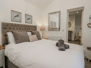 A bedroom with a large bed, a tufted headboard, two framed horse pictures, a large mirror, and a bathroom visible through an open door. - The Dove Cot (Ambleside)