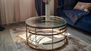Uma mesa de centro redonda de vidro, com estrutura metálica, um vaso com flores e um sofá azul ao fundo.