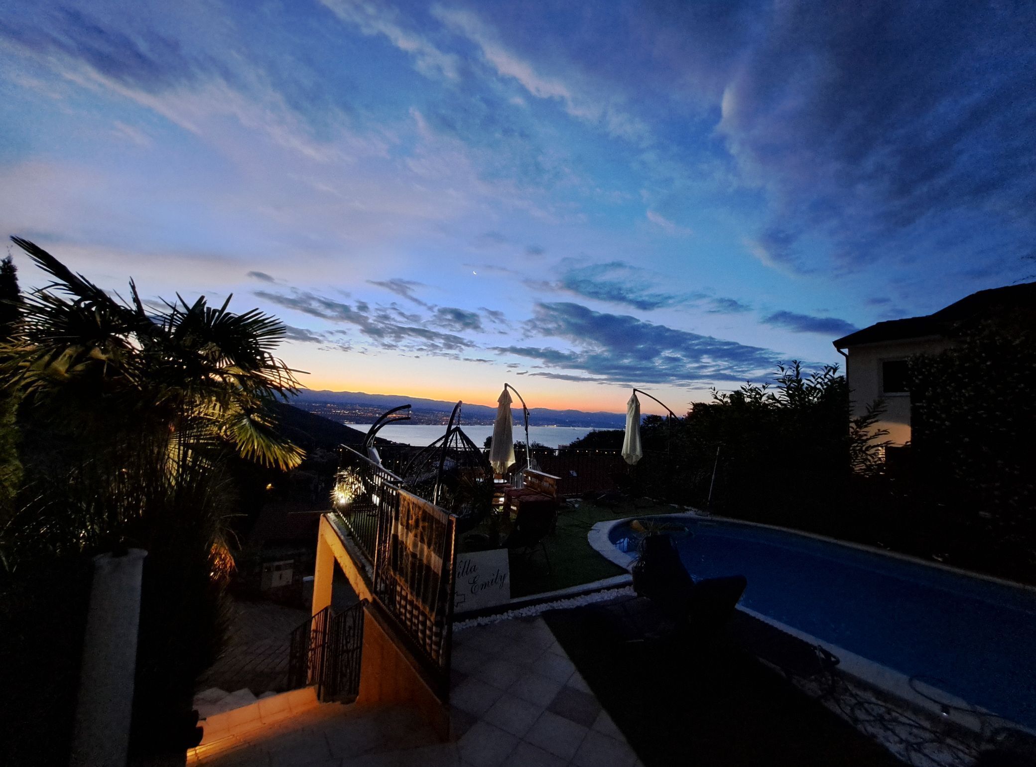 A pool area with a staircase leading to a deck, surrounded by palm trees and a view of the sunset over a body of water.