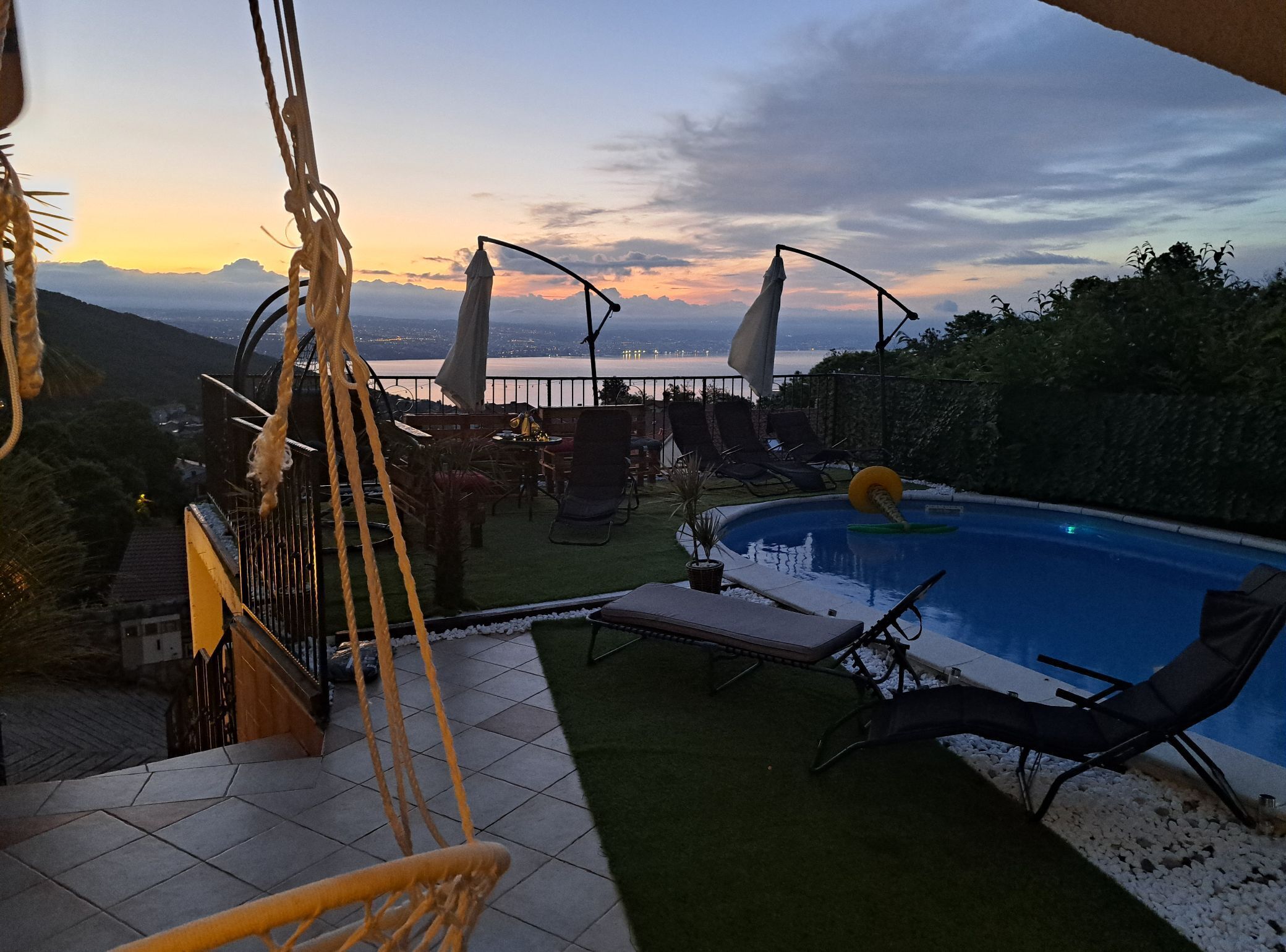 Un espace piscine avec des chaises longues, une balançoire et une vue sur un plan d’eau au coucher du soleil.