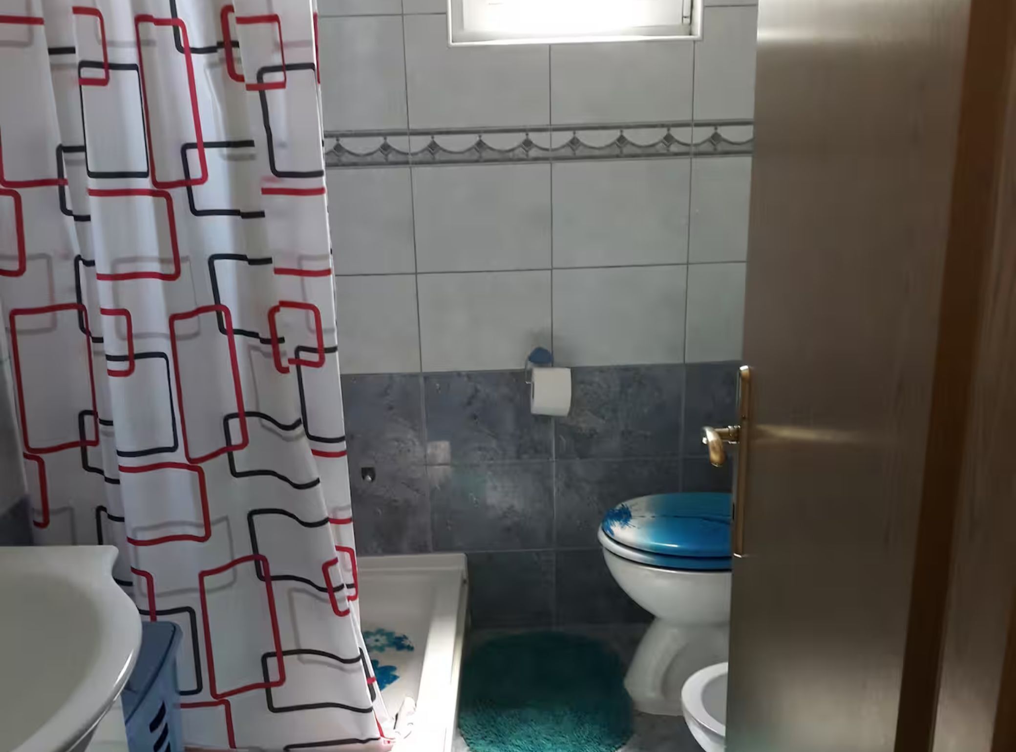 Un bagno con un WC bianco, uno sgabello del water blu, una vasca da bagno bianca e una tenda da doccia con motivo geometrico.
