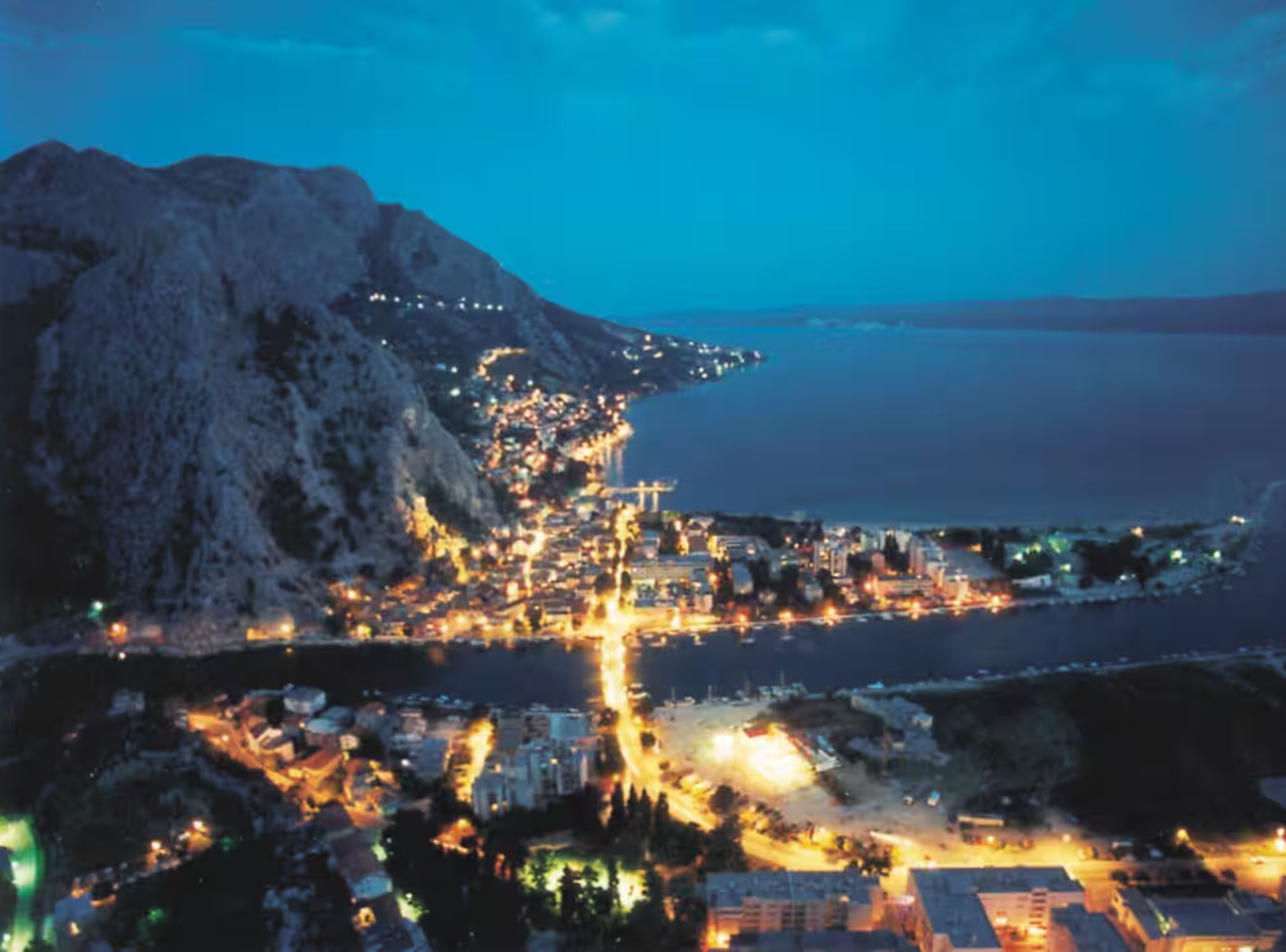 A coastal town at dusk with buildings along the waterfront and a prominent mountain in the background.