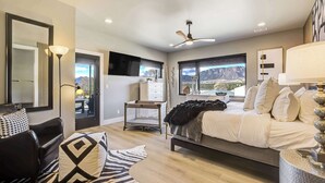 A modern bedroom with a bed, a desk, a chair, a mirror, and a view of mountains through the windows. - The Gallery! Luxury Residence w/ Front Range Views (Monument)