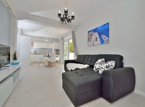 A modern living room with a dark sofa, a dining area with white chairs, and a kitchen in the background. - Villa Marija Exclusive Stay (Sukosan)