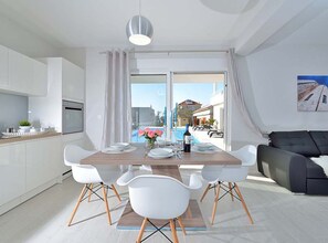A modern kitchen with white cabinets, a dining table set for four, and a view of a pool and buildings outside. - Villa Marija Exclusive Stay (Sukosan)