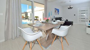 A modern dining area with a wooden table, white chairs, and a view of a pool and building outside. - Villa Marija Exclusive Stay (Sukosan)