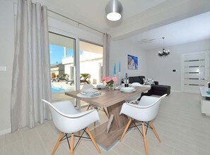 A modern dining area with a wooden table, white chairs, and a view of a pool and building outside. - Villa Marija Exclusive Stay (Sukosan)