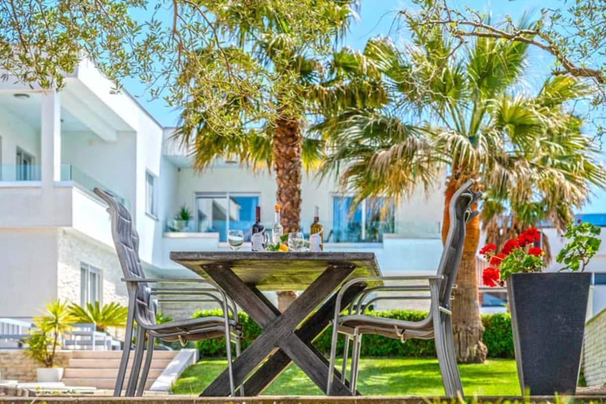 An outdoor dining area with a table and chairs, surrounded by palm trees and a building in the background.
