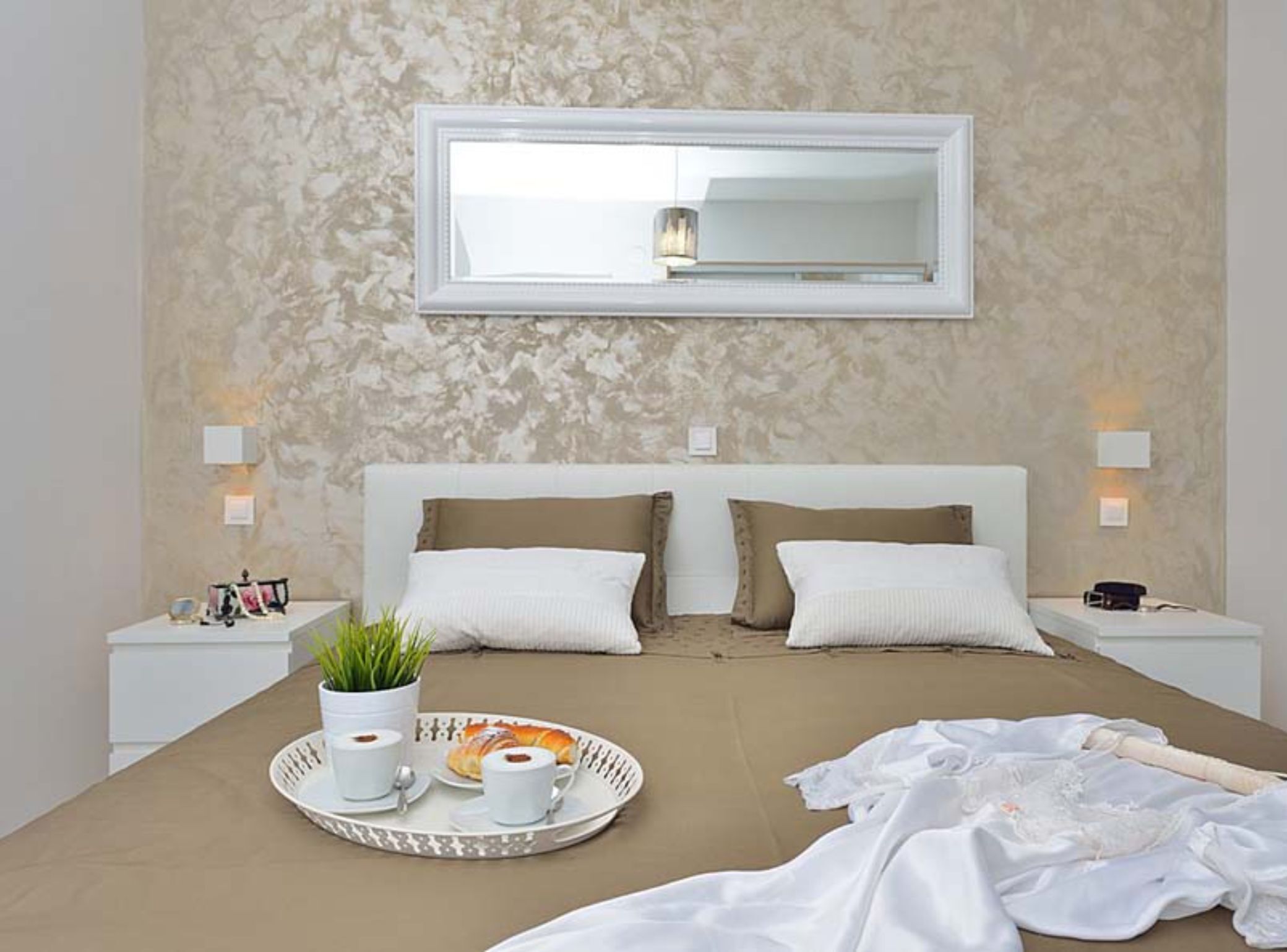 A bedroom with a large bed, a bedside table with a lamp, a tray with cups and a pastry, and a mirror above the bed.