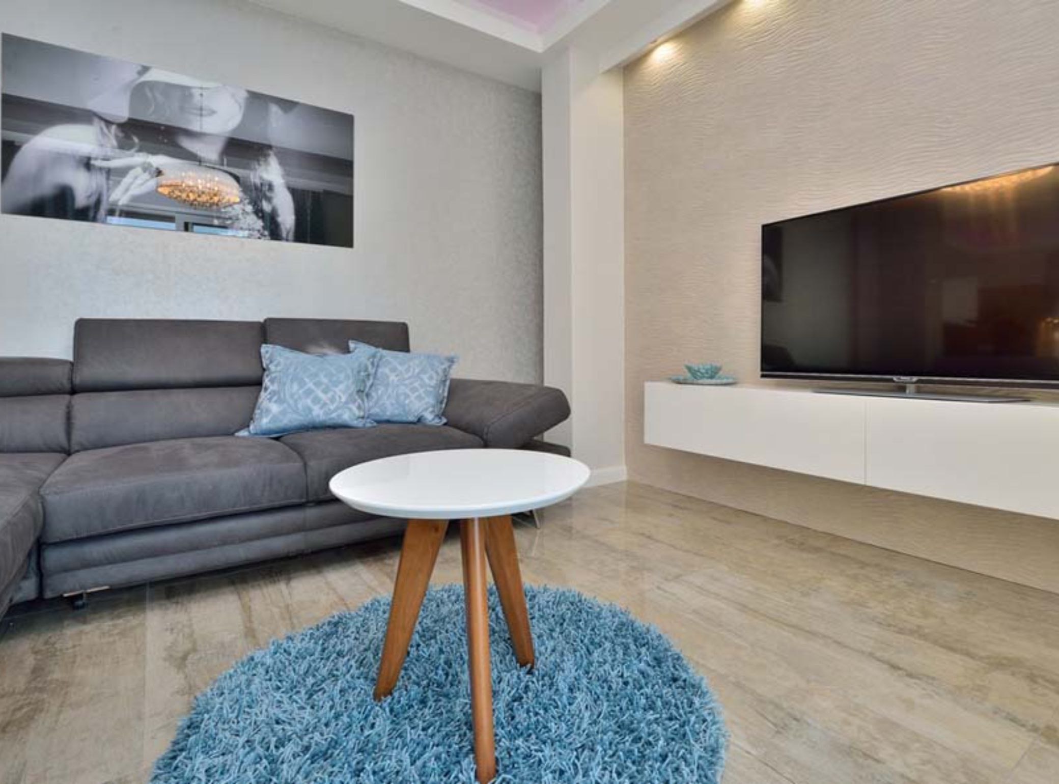 A modern living room with a grey sofa, a round white coffee table, a blue shag rug, a mounted flat-screen TV, and a wall-mounted shelf.