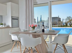 A modern kitchen with white cabinets, a dining table set for four, and a view of a swimming pool. - Villa Marija Exclusive Stay (Sukosan)