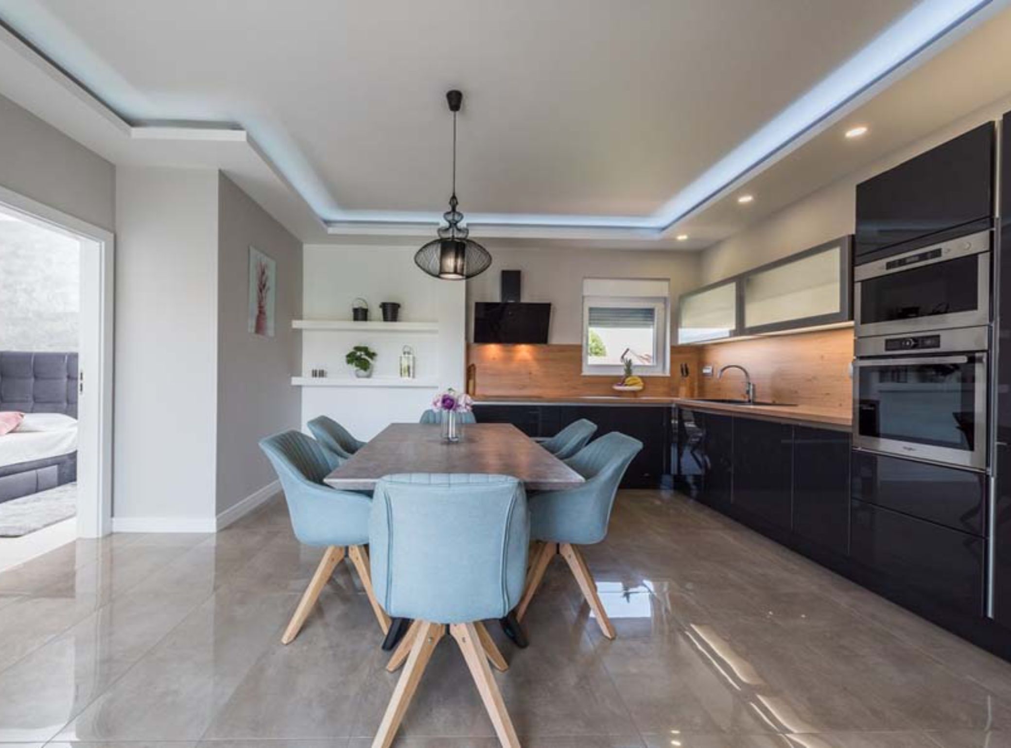 A modern kitchen with built-in appliances, a dining area with a wooden table and blue chairs, and a large window providing natural light.