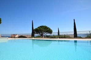 A clear blue swimming pool with a metal fence, surrounded by trees and a view of the sea. - 200 meters from the beach in Sainte-Maxime, air-conditioned apartment with ocean views and a pool (Sainte-Maxime)