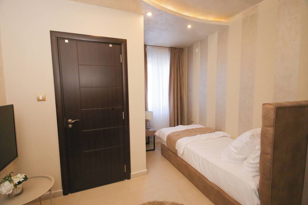 A hotel room with a dark wooden door, a bed with white bedding, a small bedside table, and a TV on the left.