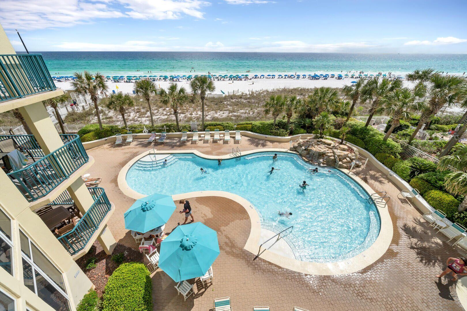 Un espace piscine avec vue sur la plage, des chaises longues, des parasols et une piscine où des personnes se baignent.
