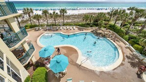 Un espace piscine avec vue sur la plage, des chaises longues, des parasols et une piscine où des personnes se baignent.