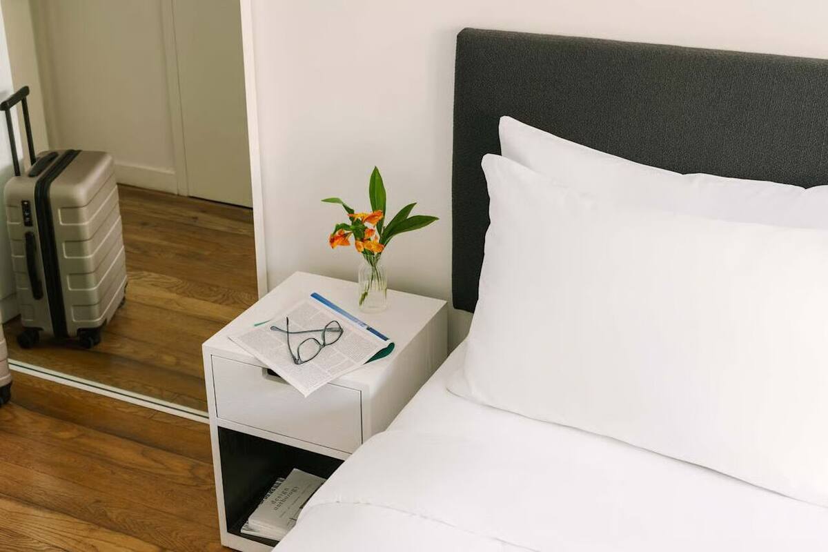 A hotel room with a bed, a nightstand with a vase of flowers, a suitcase, and a pair of glasses.