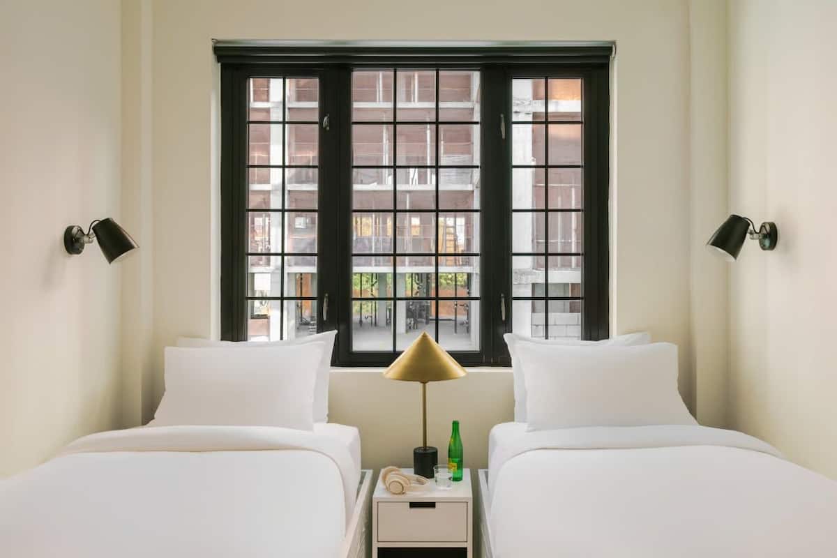 A hotel room with two beds, a window with black frames, and a nightstand with a lamp and a bottle.