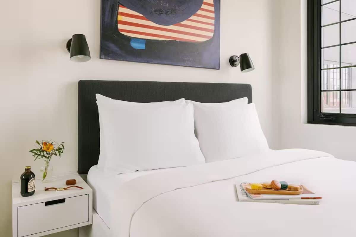 A neatly made bed with white bedding, a small bedside table with a bottle and a vase of flowers, and a wall-mounted abstract painting.