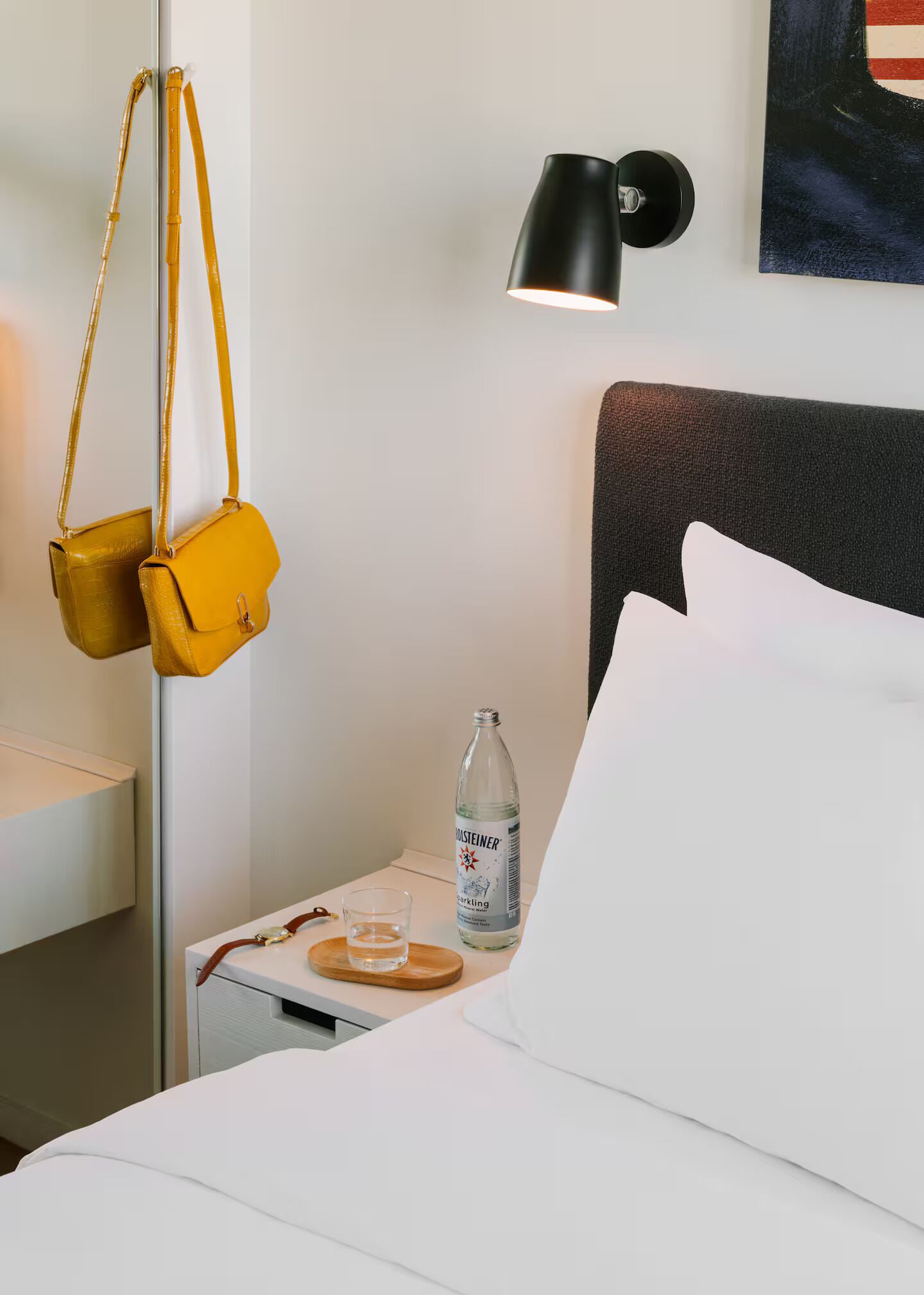 A neatly made bed with white linens, a black headboard, a bedside table with a bottle of water, a glass, and a pair of yellow bags hanging on the wall.