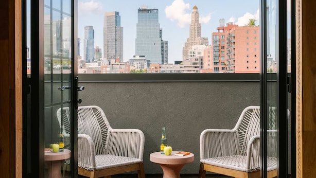 A balcony with two wicker chairs, a small round table, and a cityscape view. - The Industrialist Hotel Brooklyn (Brooklyn)
