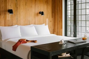 A hotel room with a wooden headboard, a bed with white linens, a desk with a laptop, a guitar, and a bottle of orange juice.