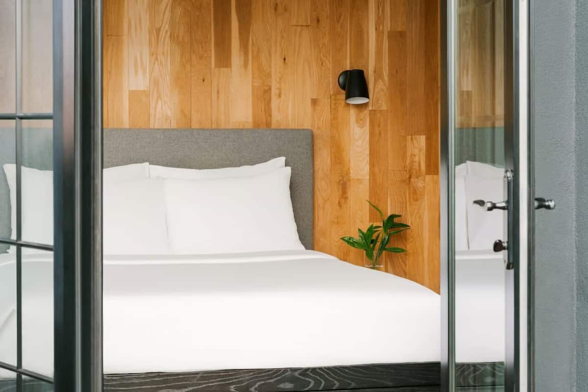 A modern bedroom with a wooden wall, a grey headboard, white bedding, a small plant, and a black wall-mounted lamp.