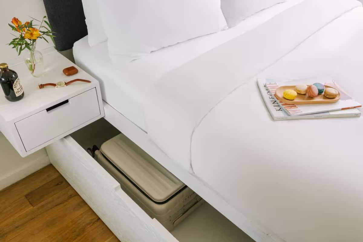A neatly made bed with a magazine and macarons on the bed. There is a small table with a bottle, a vase with flowers, and a candle.