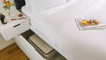 A neatly made bed with a magazine and macarons on the bed. There is a small table with a bottle, a vase with flowers, and a candle.