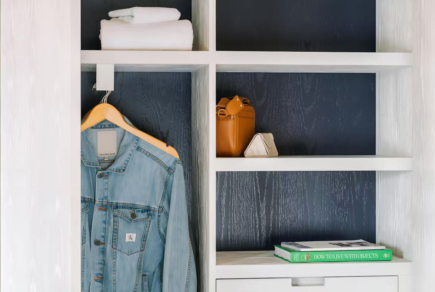 Una habitació amb una jaqueta de teixit blau sobre un penjallum, una bossa de cuir marró i un llibre anomenat 'HOW TO LIVE WITH OBJECTS' a l'estant.