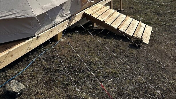 A tent with a wooden platform attached, set against a backdrop of a clear sky and distant icebergs. - Nomad Greenland (Saqqaq)