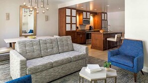 A modern living room with a grey sofa, a blue armchair, and a small table with a plant. A kitchen with wooden cabinets and a microwave is visible in the background. - 2 Bedroom, Sleep 8, Marriott Lakeshore Reserve, Spring Break (Orlando)