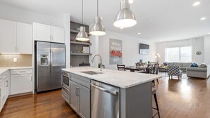 A modern kitchen with stainless steel appliances, a central island, pendant lighting, and a dining area with a gray sofa and patterned armchair. - Private Rooftop | Modern Downtown + Large Space (Nashville)
