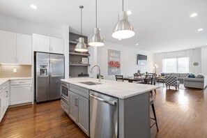 A modern kitchen with stainless steel appliances, a central island, pendant lighting, and a dining area with a gray sofa and patterned armchair. - Private Rooftop | Modern Downtown + Large Space (Nashville)